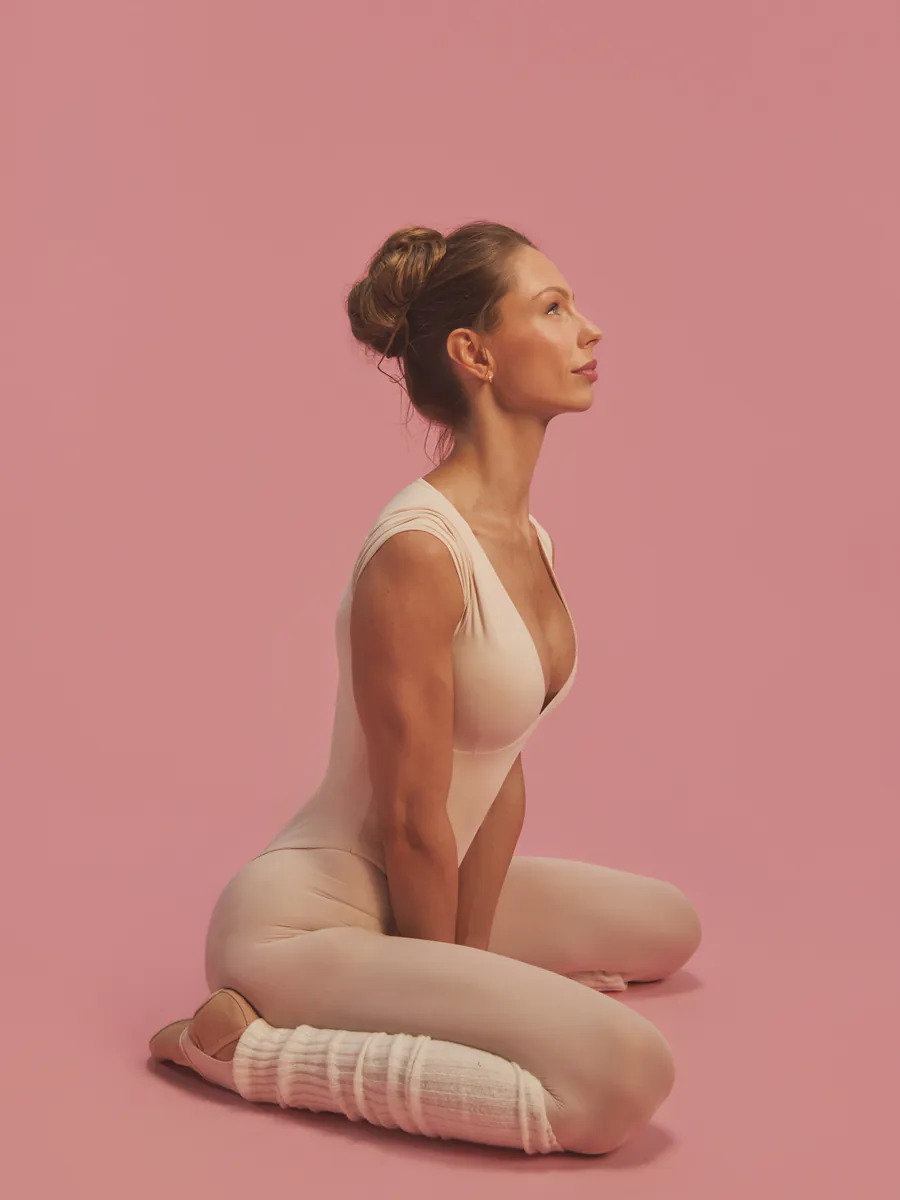 Jules kneeling in profile in a blush bodysuit and leg warmers