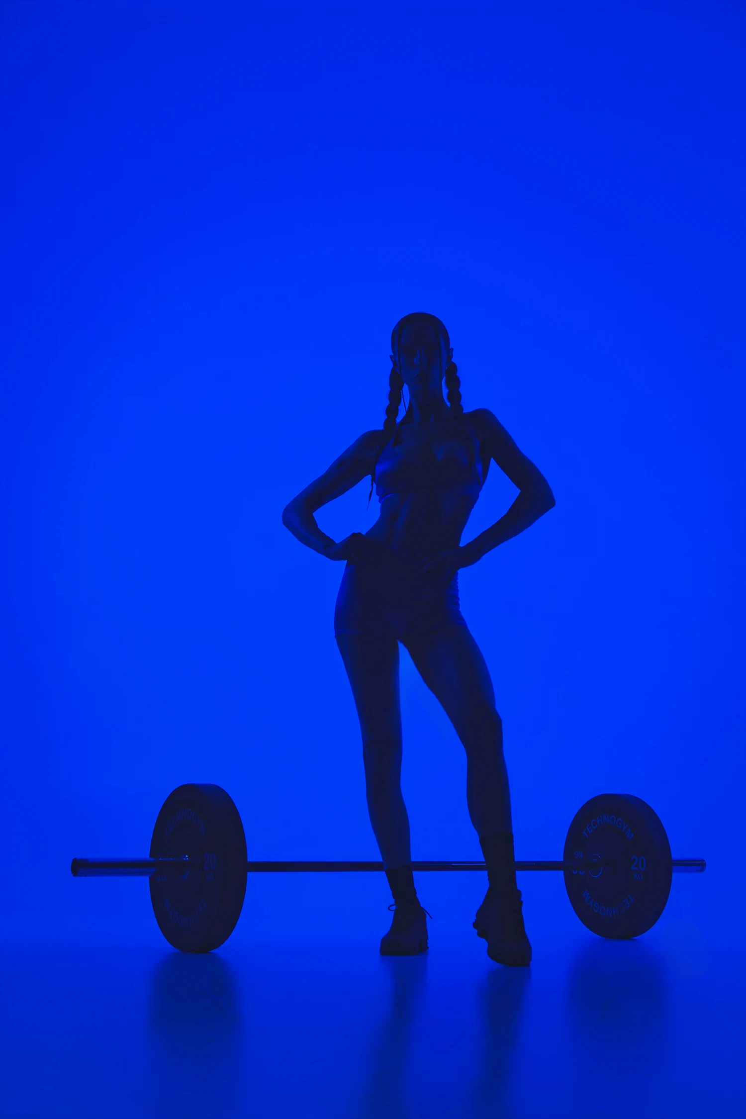 Coach Jules in silhouette, hands on hips behind a loaded barbell, against a cobalt studio backdrop