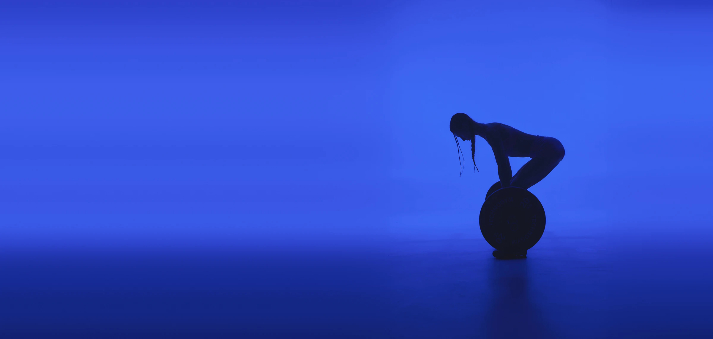 Jules in silhouette folded over a weight plate in a blue studio