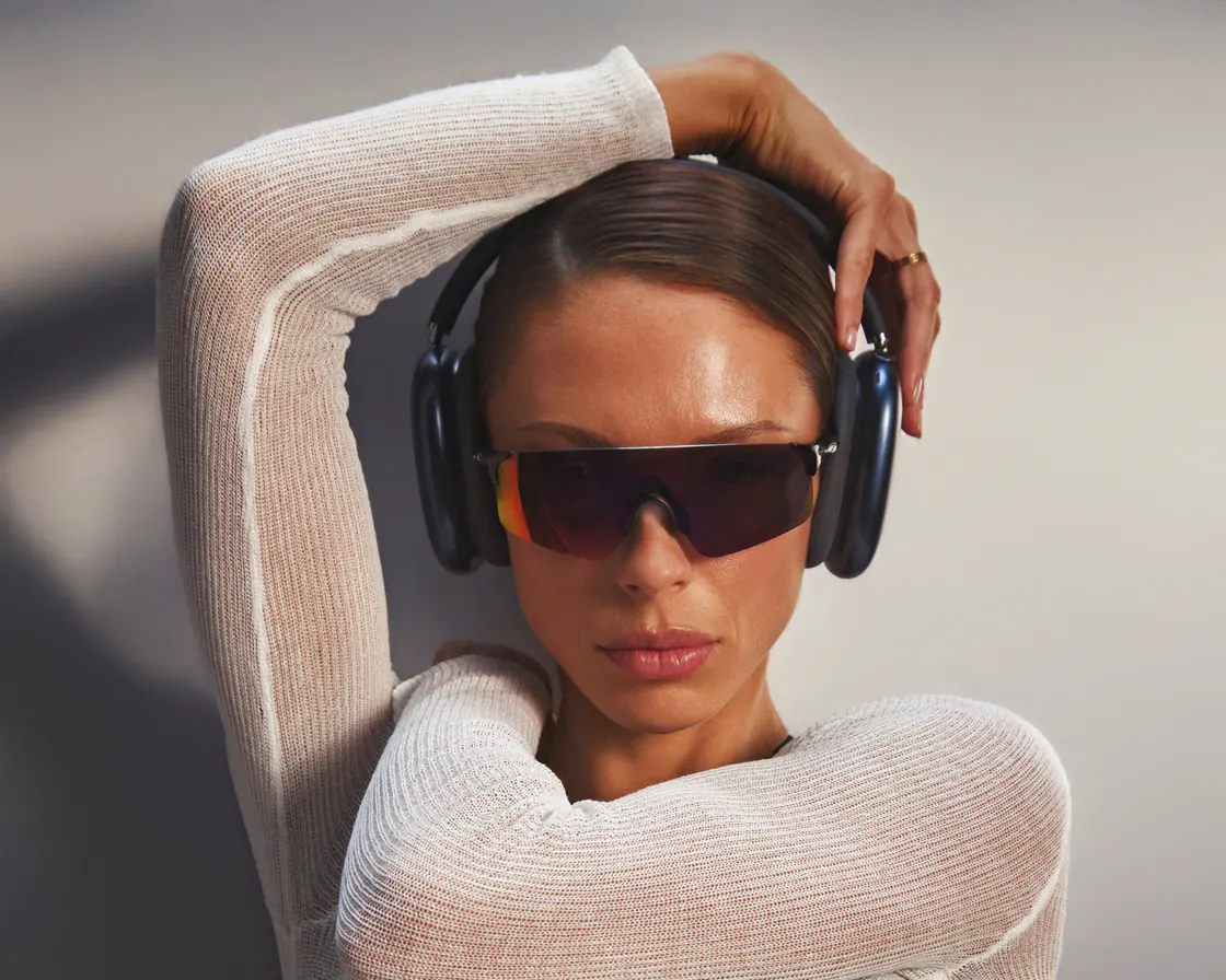 Jules in a sheer white knit top and headphones, one arm raised over her head